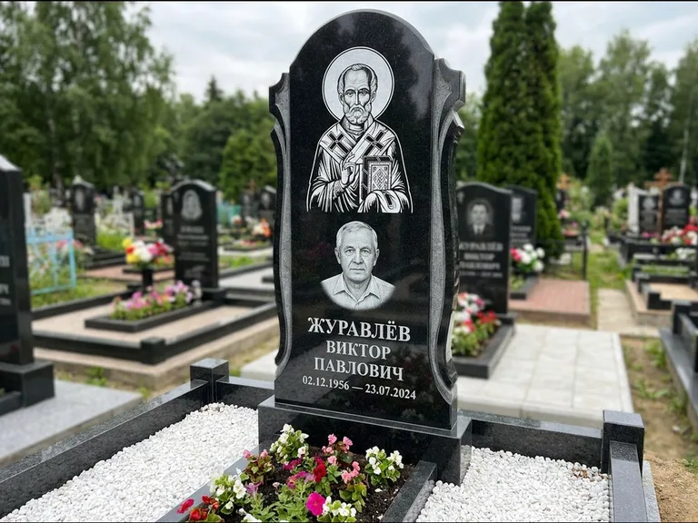 Памятник с гравировкой иконы: стела с образом в полный рост на обороте — гравировка образа Матроны Московской, низкие кусты спиреи в цвету, работа «Лебедева» в Новом Уренгое